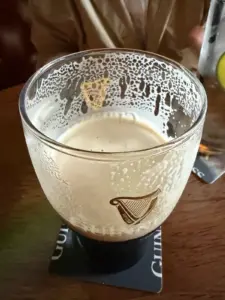 A nearly empty pint glass of Guinness sits on a coaster, leaving foam lacing on the inside. A person in a tan jacket holds a drink in the background.