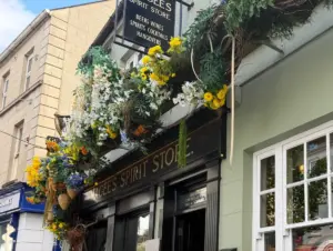 A storefront with a black sign reading “Spirit Store” is decorated with vibrant yellow, white, and green flowers and foliage above the entrance. The building is light green with a floral display hanging over the doorway.