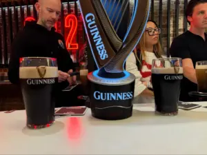 Two pints of Guinness on a bar counter, with three people sitting in the background. A prominent Guinness tap is in the center with neon lighting.