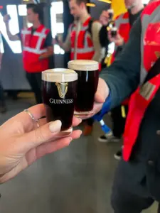 Two people hold up tiny glasses of Guinness in a toast. Both wear red safety vests, and other people in similar vests are visible in the blurred background.