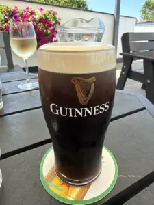 A pint of Guinness stout on a table outdoors, with a wine glass, water jug, and flowers in the background. The beer sits on a coaster, and sunlight brightens the setting.