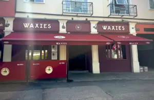 Street view of a maroon and cream storefront named “Waxies,” with signs, awnings, and barriers displaying the name. Two balconies with railings are above the shop. Sunlight creates a streak across the image.