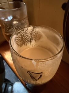 A half-finished pint of Guinness with foam marks on the glass sits beside a glass of water with ice on a wooden surface in warm lighting.
