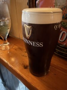 A pint glass of Guinness stout with a creamy head sits on a wooden bar next to a glass of water. The Guinness logo and harp symbol are visible on the glass.