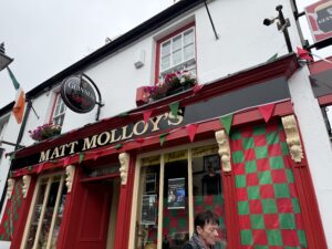 The exterior of Matt Molloy’s pub, decorated with red and green checkered flags, bunting, and flower boxes. The sign for Guinness is visible above the entrance. A person walks past the front.