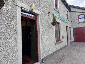 A textured gray building with red-framed windows and doors, green hanging signs, a hanging plant basket, and an open door leading inside—this is O’Neill’s in Letterbarra. The concrete path runs alongside the building.