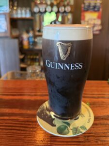 A pint of Guinness stout with a creamy head sits on a coaster atop a wooden bar at O’Neill’s in Letterbarra, with a blurred pub background and beer taps visible behind it.