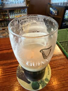 A half-finished pint of Guinness sits on a coaster atop a wooden bar at O’Neill’s in Letterbarra, with foam clinging to the inside of the glass. Shelves with glassware and bottles are visible in the background.