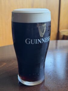 A pint glass of Guinness stout with a creamy head sits on a wooden table, sunlight shining in the background.
