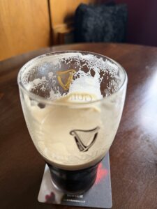 A half-finished pint of Guinness sits on a coaster atop a wooden table, with foam clinging to the inside of the glass. A dark cushion is in the blurred background.