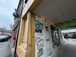 A narrow street with a yellow building featuring vintage signs and a white door. String lights hang above, and the sidewalk is partially covered with a checkered green and yellow awning.