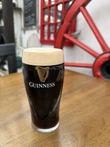 A pint of Guinness stout sits on a wooden table. In the background, there is part of a red wagon wheel and a chalkboard menu visible.
