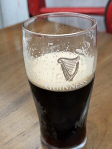 A half-full pint glass of Guinness stout sits on a wooden table. The creamy head leaves lacing on the glass, and the iconic Guinness harp logo is visible on the front. A red object is blurred in the background.