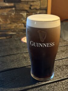 A pint of Guinness stout with a creamy head sits on a dark wooden table, with a blurred stone wall and coaster in the background.