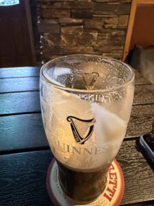A partially finished pint of Guinness sits on a coaster atop a dark wooden table, with foam clinging to the inside of the glass. Stone wall and door are visible in the background.