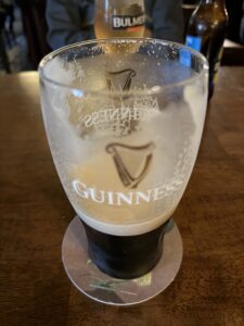 A partially finished pint of Guinness sits on a coaster on a wooden table, with foam lacing the glass. Other drinks and blurred background elements are visible behind it.