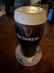 A pint of Guinness stout with a creamy head sits on a coaster on a wooden table. Other drinks are visible in the background, with a dim, cozy pub atmosphere.