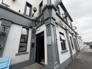 A wide-angle view of the gray and white exterior of a traditional pub with open windows, hanging lights, and a sign above the door, taken on a cloudy day.