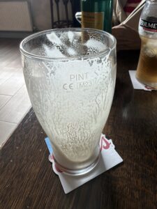 An empty pint glass with foam residue sits on a coaster on a wooden table, next to a bottle and a glass of Bulmers cider. A person’s arm is partially visible in the background.