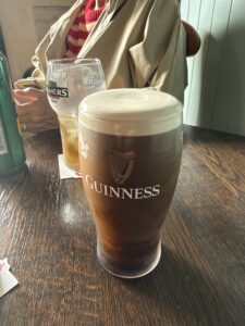 A pint of Guinness on a wooden table, with a partially filled Bulmers glass and a person in a tan coat and red-striped shirt blurred in the background.